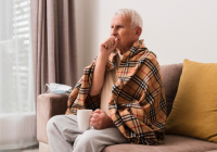 Homem idoso, envolvido por uma manta, com o punho direito cobrindo a boca, indicando doença respiratória.