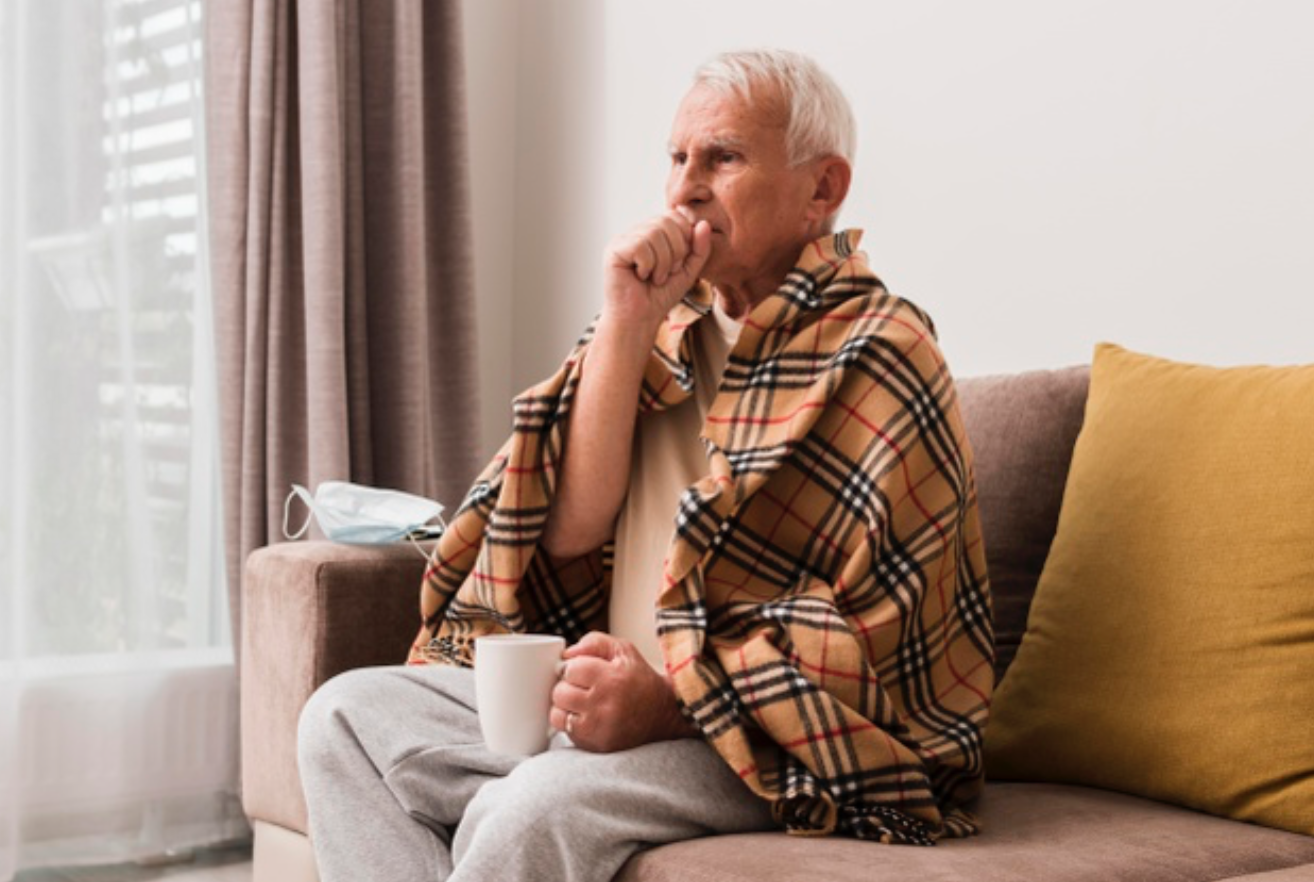 Homem idoso, envolvido por uma manta, com o punho direito cobrindo a boca, indicando doença respiratória.