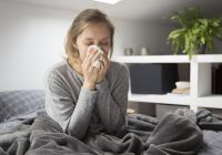 Sick young Caucasian woman covered with grey blanket sitting on bed with closed eyes, blowing nose with napkin. Illness, pain concept